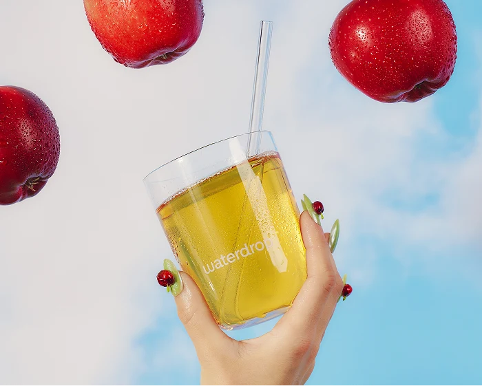 Une main tient un verre de jus jaune avec une paille, entouré de pommes rouges et d'un ciel bleu.