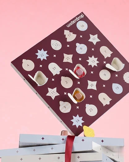 Calendrier de l'Avent avec des portes blanches numérotées sur un carré violet, au-dessus de cadeaux emballés sur fond rose.