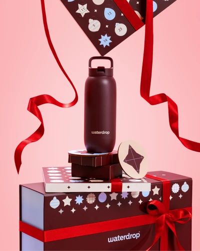 Bouteille d'eau noire avec bouchon et poignée sur des coffrets-cadeaux empilés, noués de rubans rouges sur fond rose.