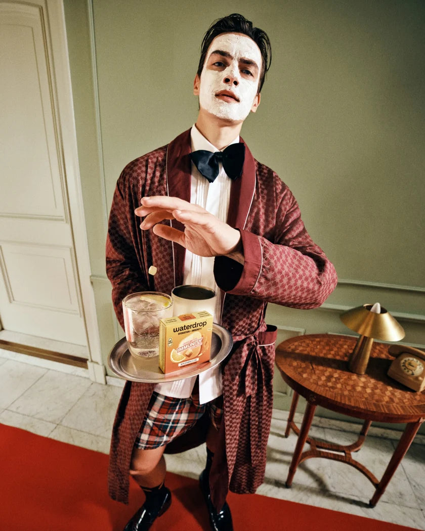 Personne au visage maquillé en blanc, portant un peignoir bordeaux et une cravate papillon, tient un plateau avec deux verres.