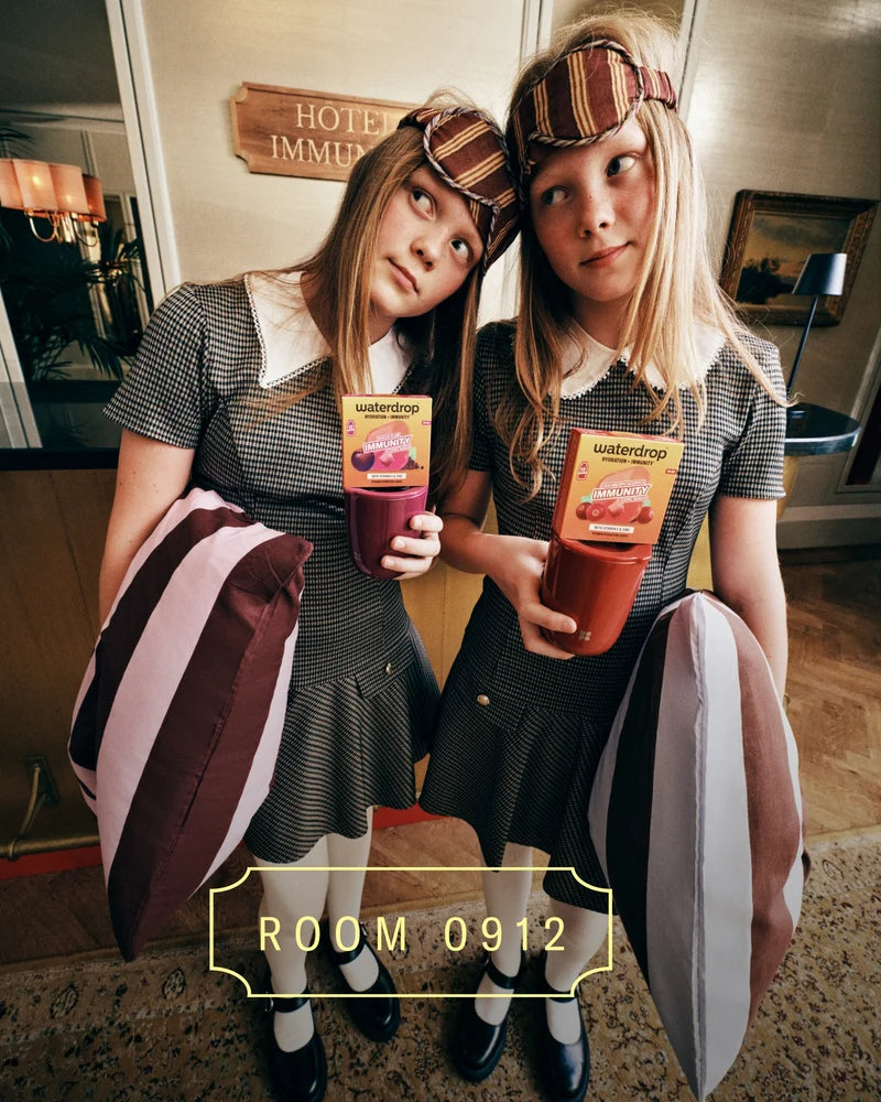 Deux filles en tenues assorties avec des bandeaux à carreaux tiennent des boissons colorées dans le hall d’un hôtel, debout côte à côte avec des sacs rayés.