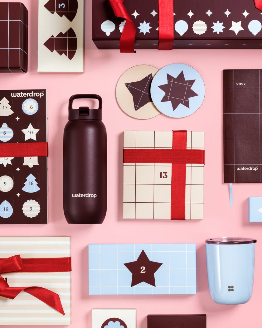 Calendriers de l'Avent et boîtes cadeaux à rubans rouges sur fond rose, avec une bouteille noire et une tasse bleu clair.