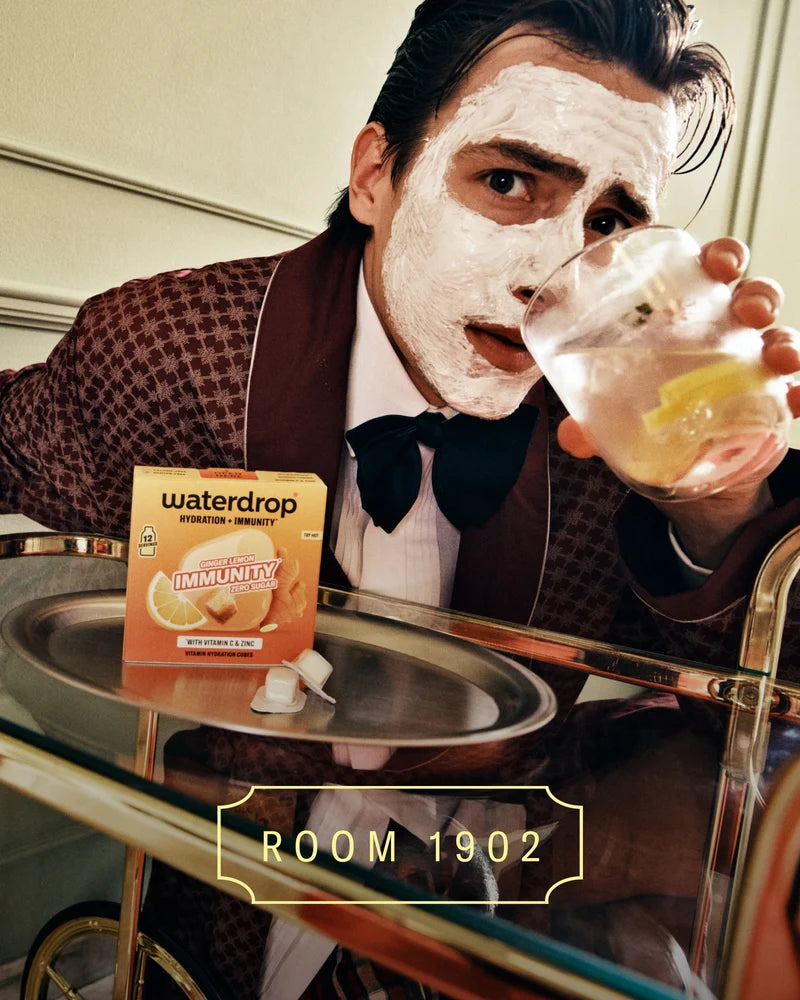 Personne en costume marron à carreaux avec nœud papillon sombre porte un masque blanc et tient un verre de boisson à côté d’un plateau avec un produit en boîte.